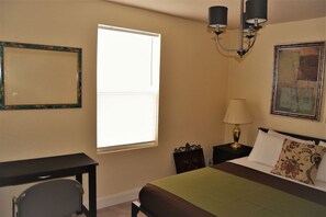 A bedroom with a bed, a desk, a chair, a window with blinds, a nightstand with a lamp, and a framed picture on the wall. - The Gateway: experience a stylish and welcoming lodging, perfect for groups. (Grand Forks)