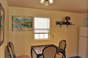Un coin repas avec une table et des chaises, une fenêtre avec des stores, une étagère murale agrémentée d’objets décoratifs et un réfrigérateur.