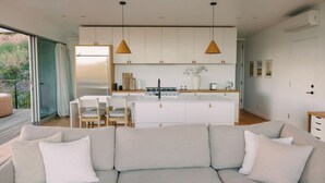 A modern living room with a sofa, a dining area with chairs, a kitchen with cabinets, pendant lights, and a sliding glass door leading to an outdoor area. - A modern light-filled home with views of the woods (Bonita)