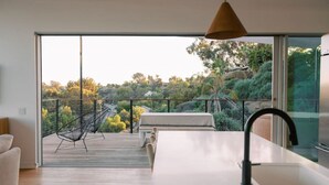 A modern kitchen with a large window, a dining area with a view, and a deck with outdoor furniture. - A modern light-filled home with views of the woods (Bonita)