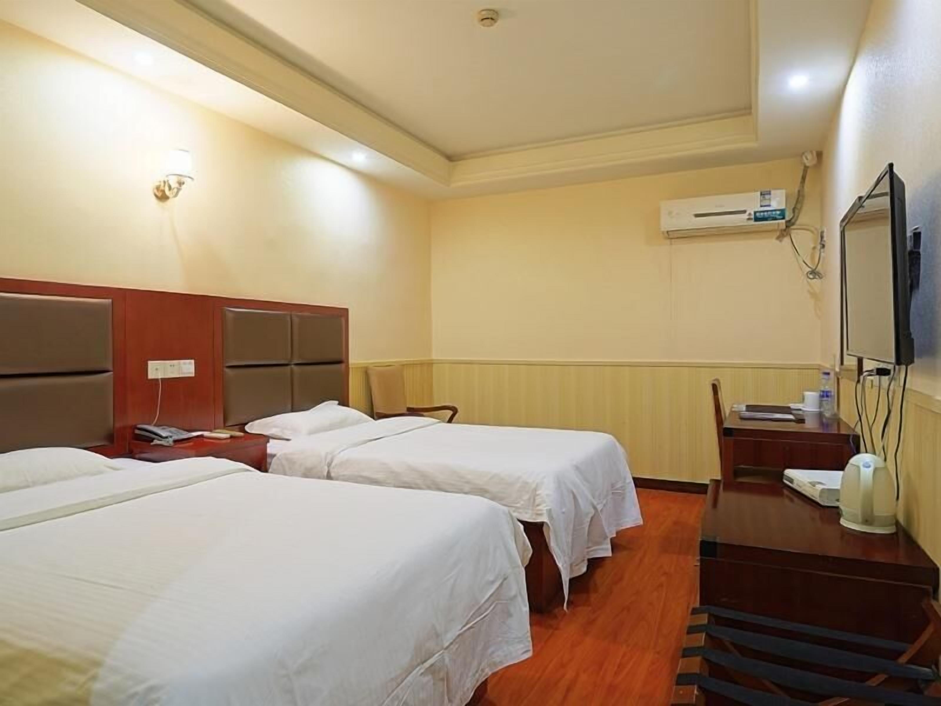 A hotel room with two single beds, a desk with a chair, a flat-screen TV, an air conditioning unit, and a wall-mounted light fixture.