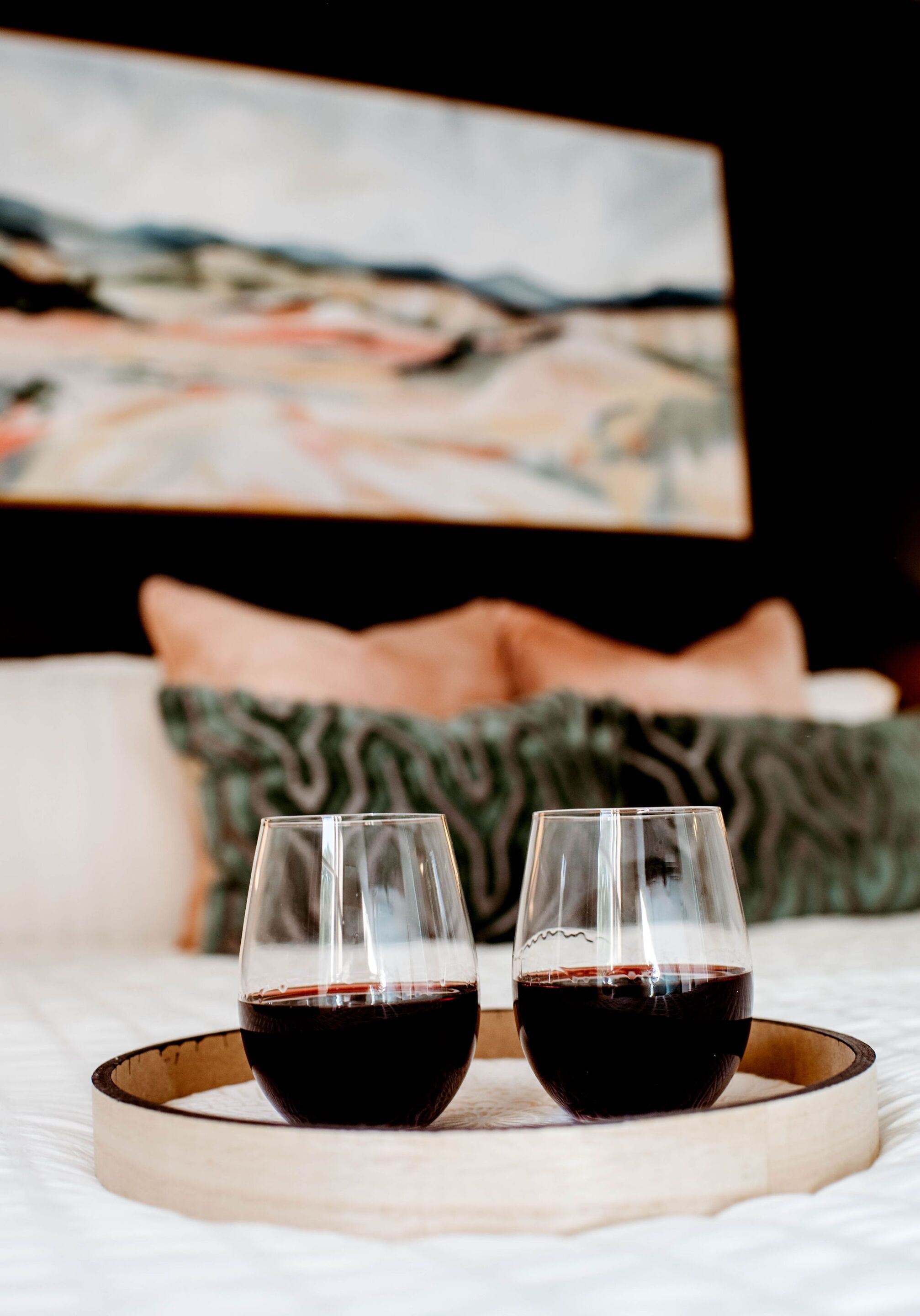 Two glasses of red wine on a tray, placed on a bed with pillows and a painting in the background.