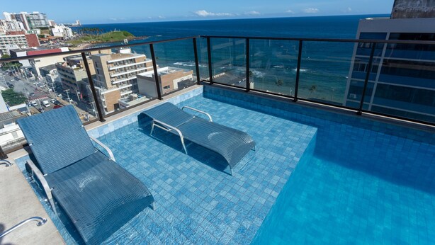 Una piscina en la azotea con dos sillones reclinables, una vista despejada del océano y un paisaje urbano al fondo.