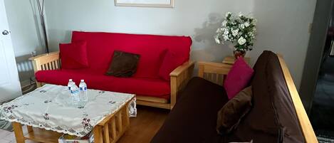 Una sala de estar con un sofá rojo, un sofá marrón, una mesa de centro de madera con un mantel de encaje, y un jarrón con flores blancas.