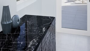 A modern bathroom with a black marble countertop, a window with a view of the sea, and a door leading to a balcony with a chair. - The Ampersand Sky Villa: Iconic, Artful Luxury (Charlotte Amalie, Saint Thomas)
