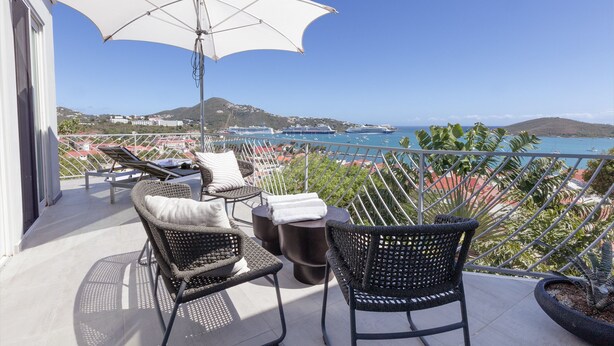 A balcony with wicker chairs, a table, and an umbrella, overlooking a coastal view with buildings and boats. - The Ampersand Sky Villa: Iconic, Artful Luxury (Charlotte Amalie, Saint Thomas)