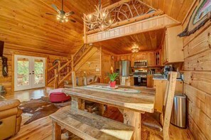 L’intérieur d’une cabane en rondins, avec une table à manger en bois, des bancs, un coin cuisine et un escalier.
