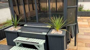 Un espacio al aire libre techado con mesa y bancos, rodeado de macetas con plantas.