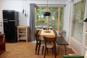 Un coin repas avec une table et des chaises, un réfrigérateur noir, une petite table en bois et une vue sur la verdure à l’extérieur.