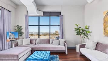 Un salon moderne avec un canapé d’angle, un pouf bleu et une grande fenêtre donnant sur la ville.