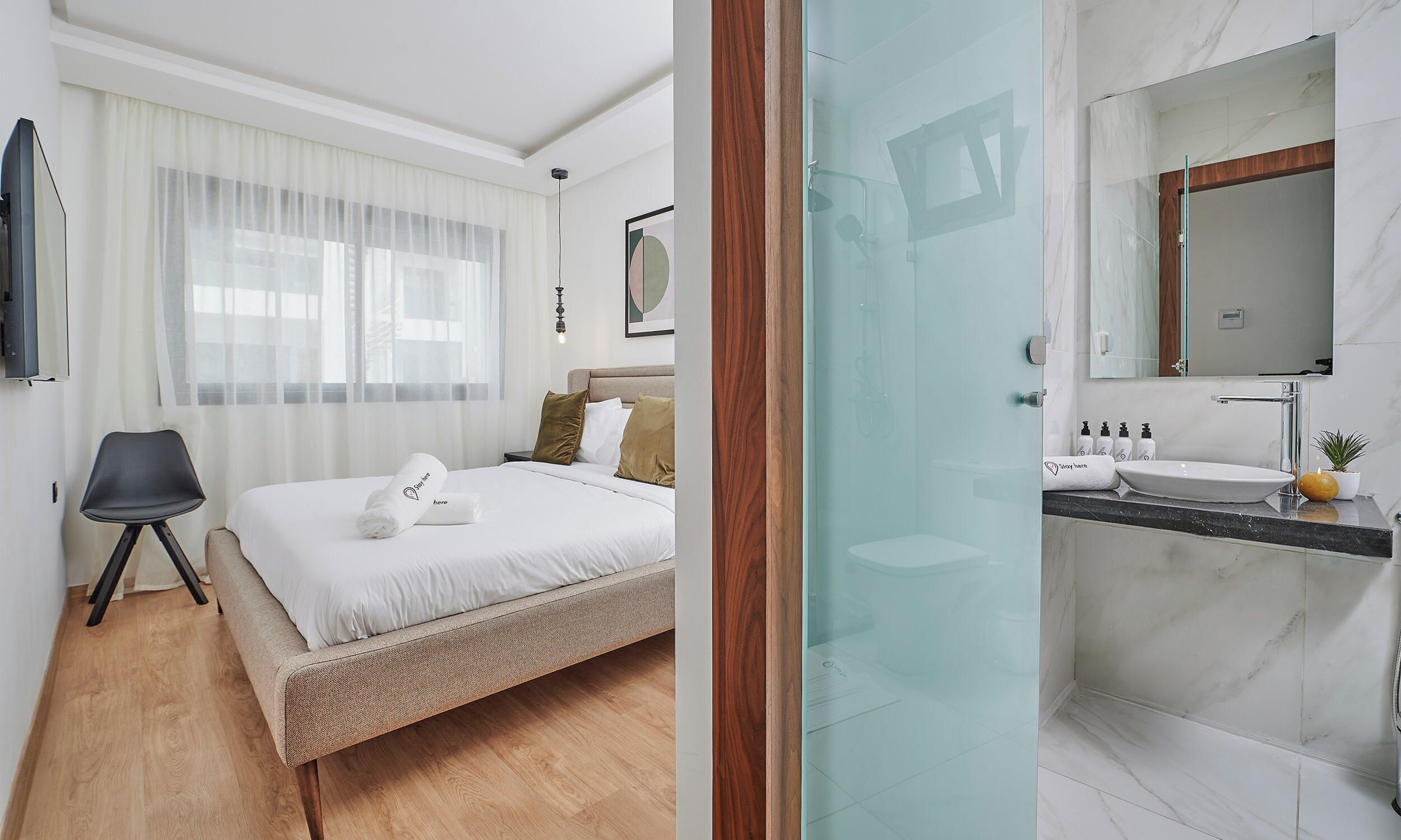Une chambre d’hôtel comprenant un lit, une chaise, une fenêtre avec des rideaux, une table de chevet et une salle de bain avec un lavabo, un miroir et des articles de toilette.
