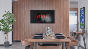 A modern dining area with a wooden table set for four, a TV mounted on the wall displaying "NETFLIX", and a potted plant to the left.