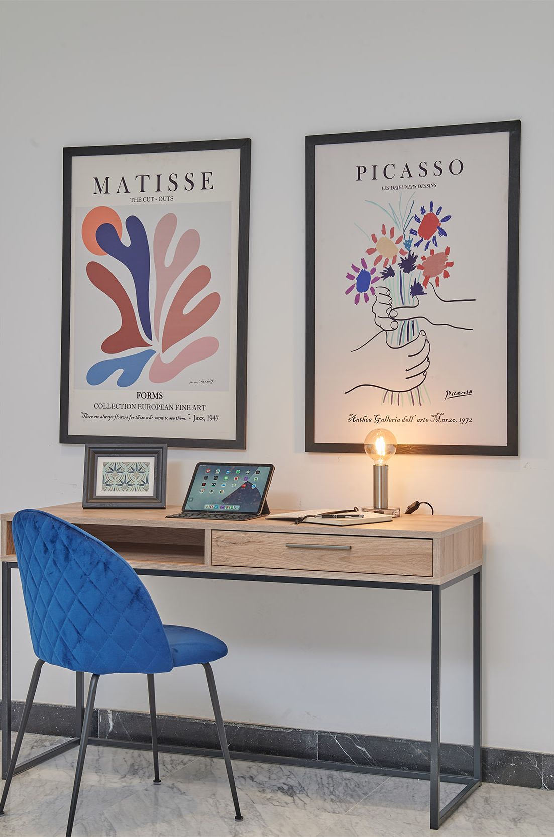 Un bureau avec un ordinateur portable, un tableau de Matisse encadré, un tableau de Picasso encadré et une chaise bleue.