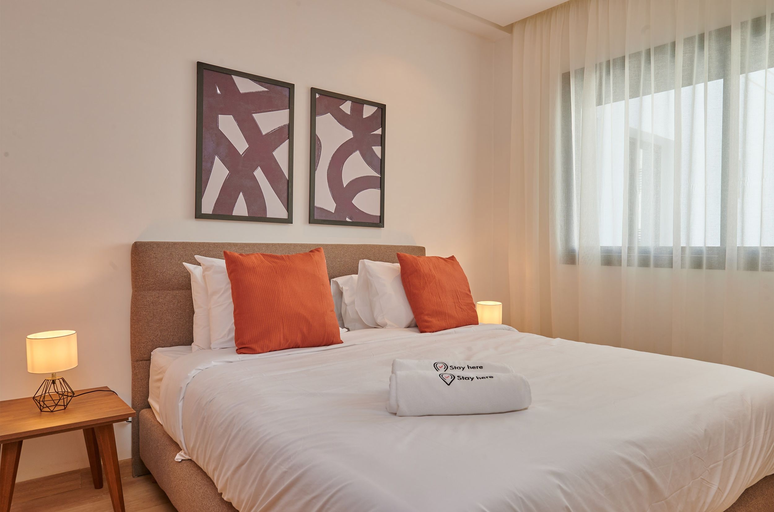 Een netjes opgemaakt bed met witte lakens en oranje kussens, een nachtkastje met een lamp, en twee abstracte kunstwerken aan de muur.