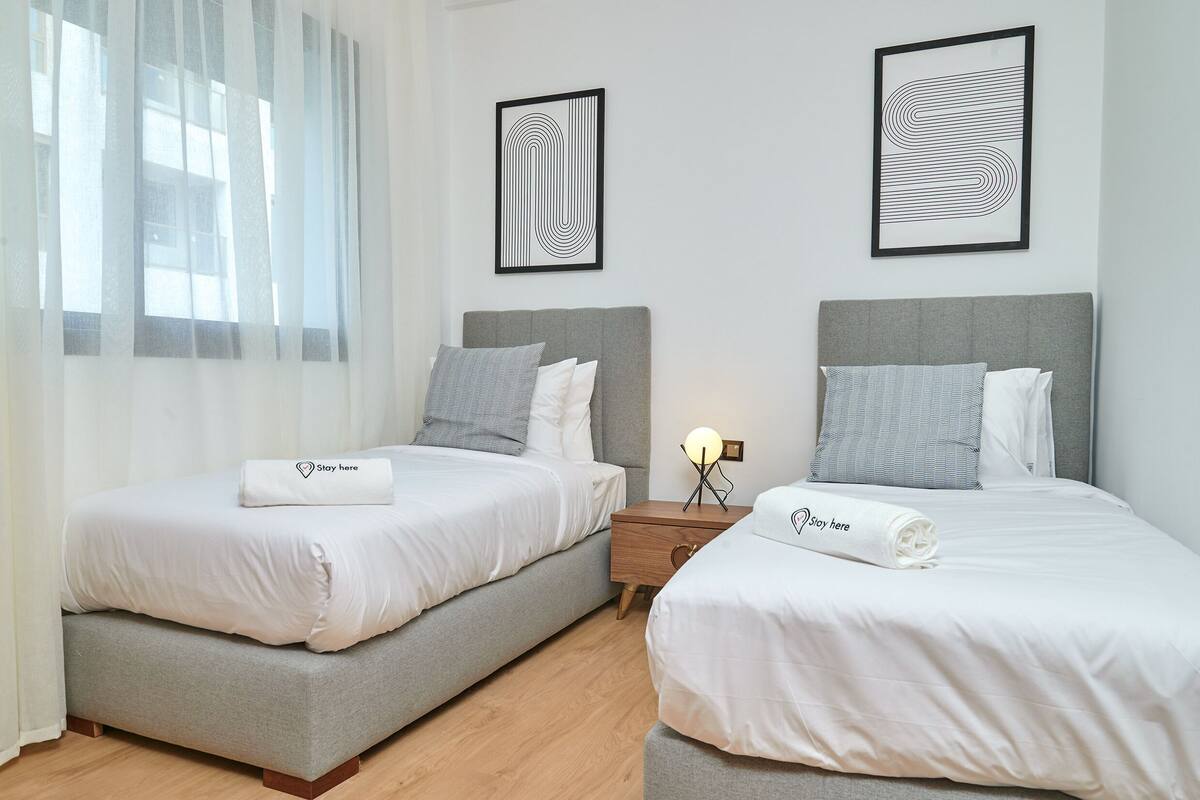 A hotel room with two beds, each with white bedding and grey headboards. There are framed abstract artworks on the walls, a bedside table with a lamp, and a window with sheer curtains.
