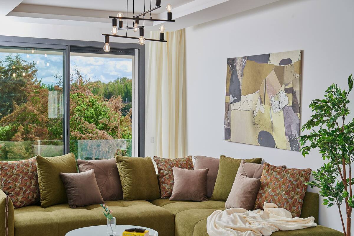 A modern living room with a green sofa, multiple cushions, a large abstract painting, and a view of trees through a large window.