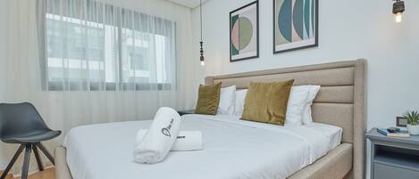A neatly made bed with a white comforter and a rolled towel, a gray nightstand with a lamp, and a chair.