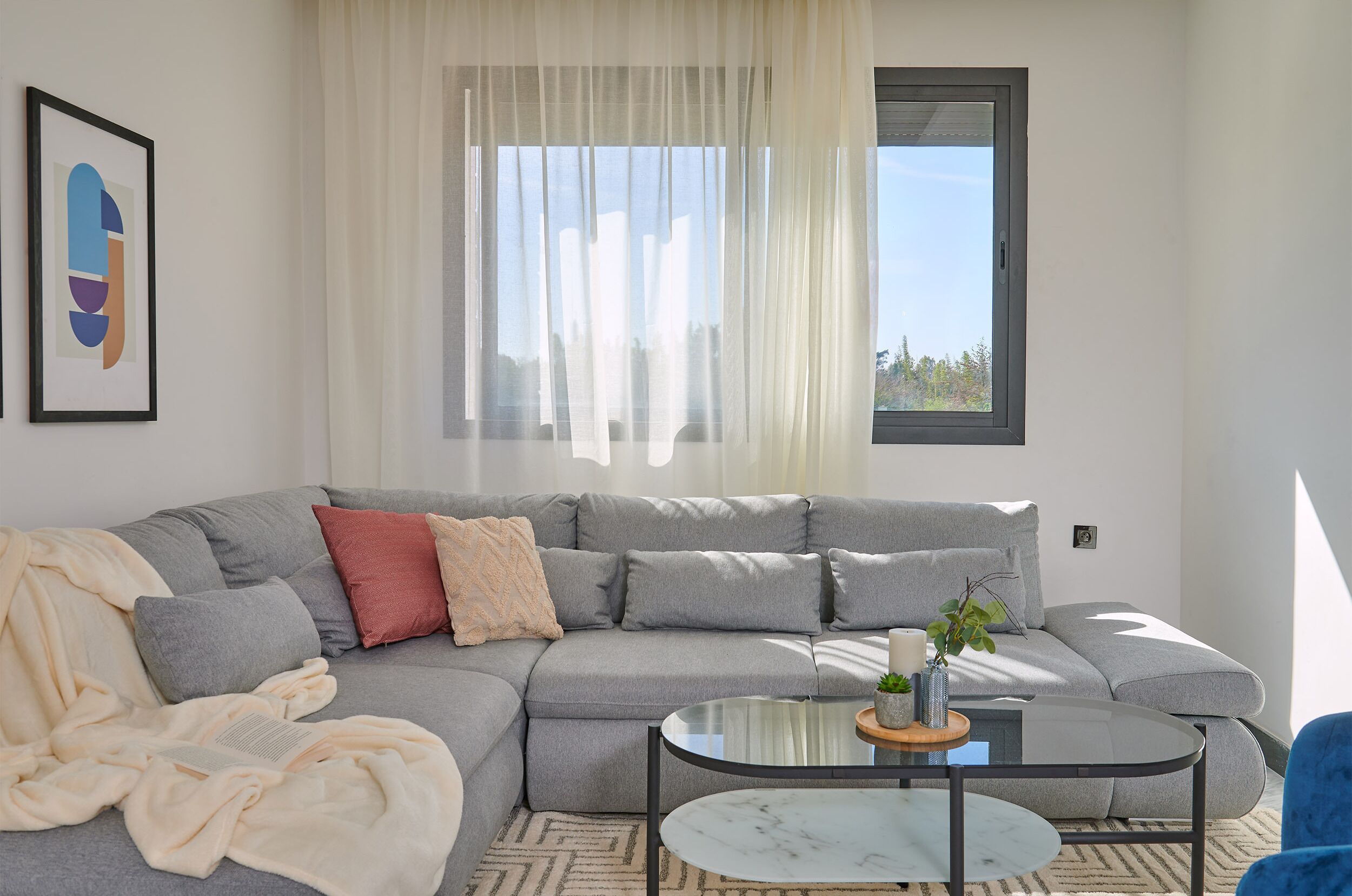 Een moderne woonkamer met een grijze modulaire bank, een glazen salontafel en uitzicht op de omgeving door een raam met transparante gordijnen.