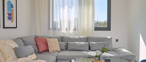 A modern living room with a grey sectional sofa, a glass coffee table, and a view of the outdoors through a window with sheer curtains.