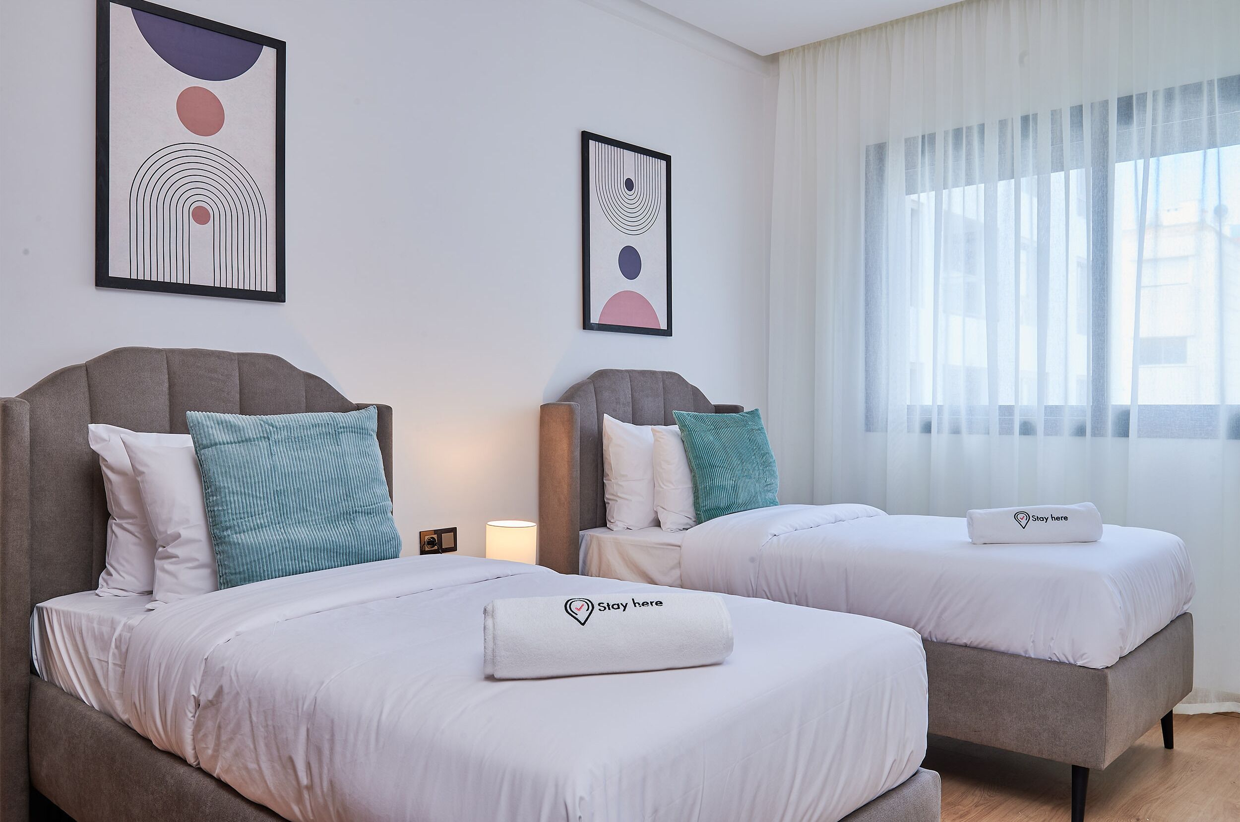 A hotel room with two beds, each with a pillow and a towel with a logo. There are two framed artworks on the wall.