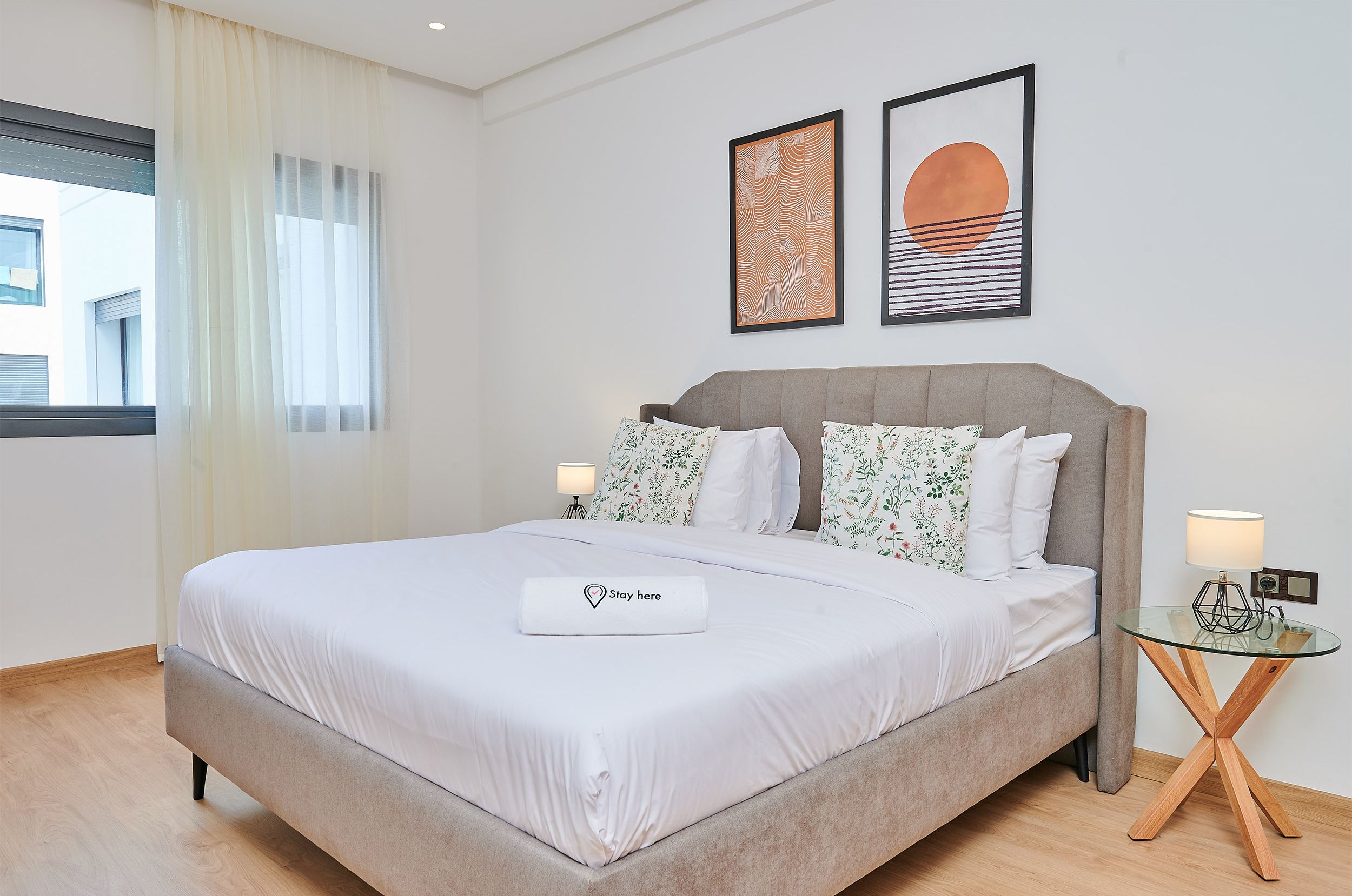 A neatly made bed with a "Stay here" pillow, a bedside lamp, and a framed artwork above it.