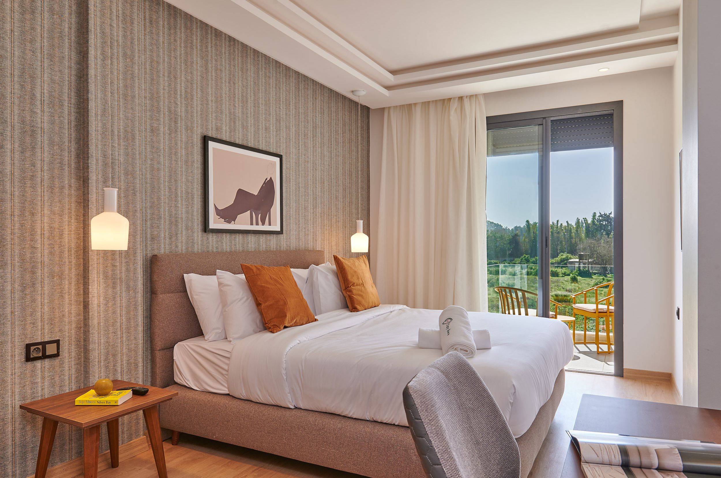 A modern hotel room with a large bed, a small table, a chair, and a balcony with a view of greenery.
