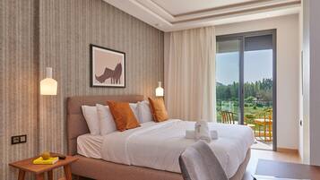 A modern hotel room with a large bed, a small table, a chair, and a balcony with a view of greenery.