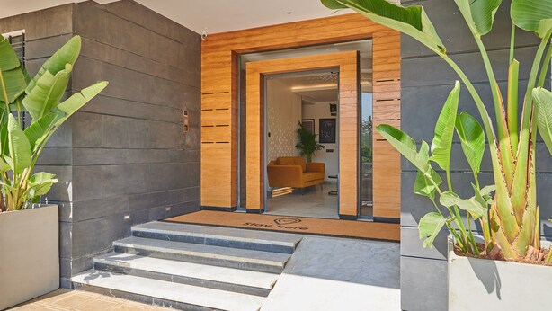 A modern building entrance with a wooden-framed door, a reception area with a sofa, and large potted plants.