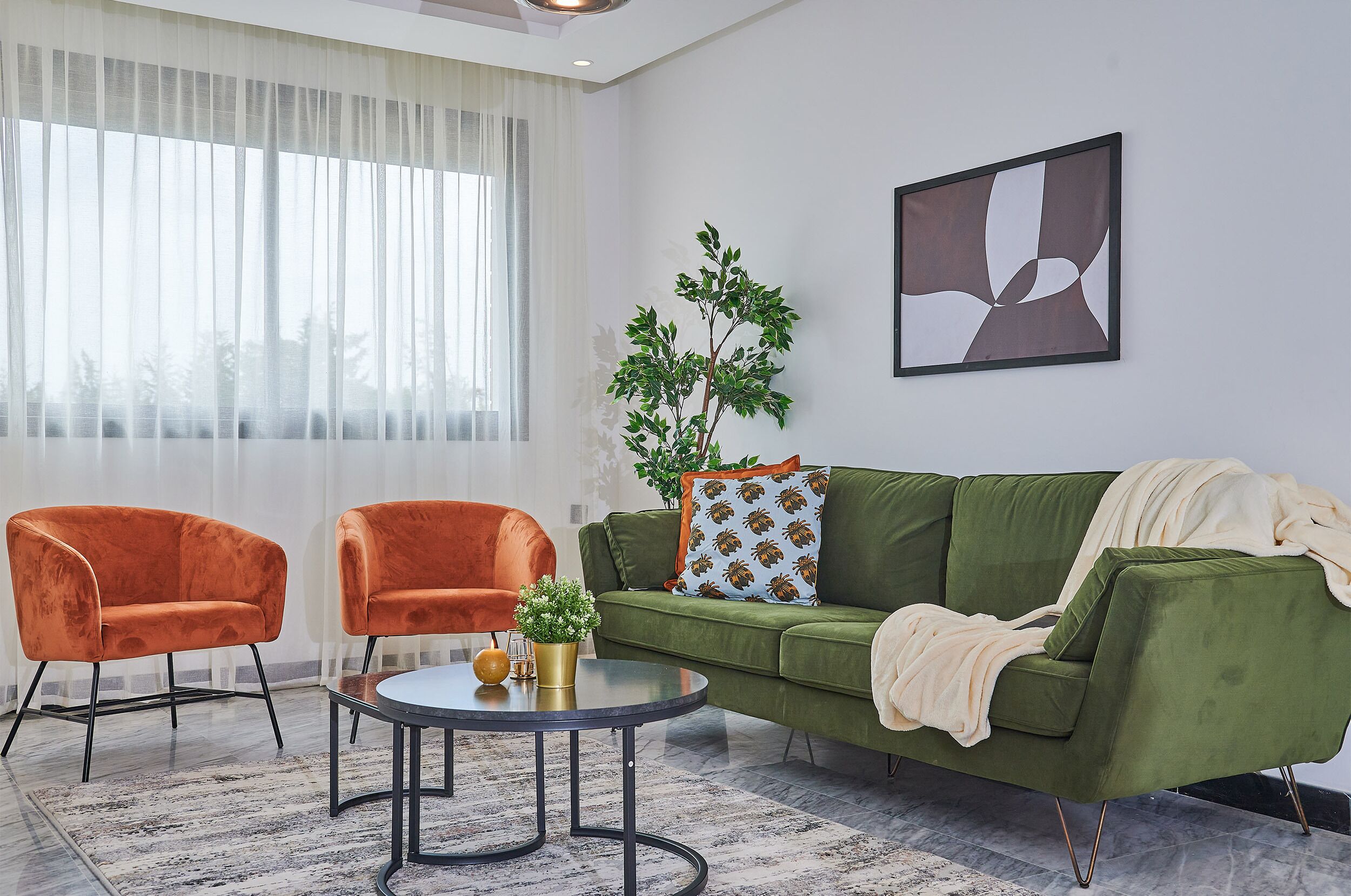 A living room with a green sofa, two orange armchairs, a round coffee table, and a large window with sheer curtains.