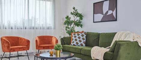 A living room with a green sofa, two orange armchairs, a round coffee table, and a large window with sheer curtains.