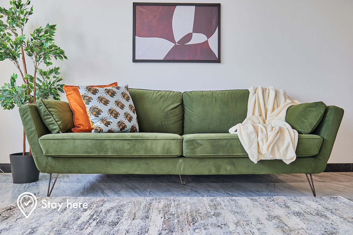 A green sofa with multiple cushions, a white throw blanket, and a potted plant to the left.