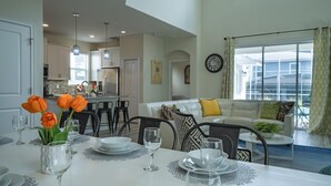 Un coin repas avec une table dressée pour quatre personnes, une cuisine en arrière-plan et un salon avec un canapé et une vue sur l’extérieur.