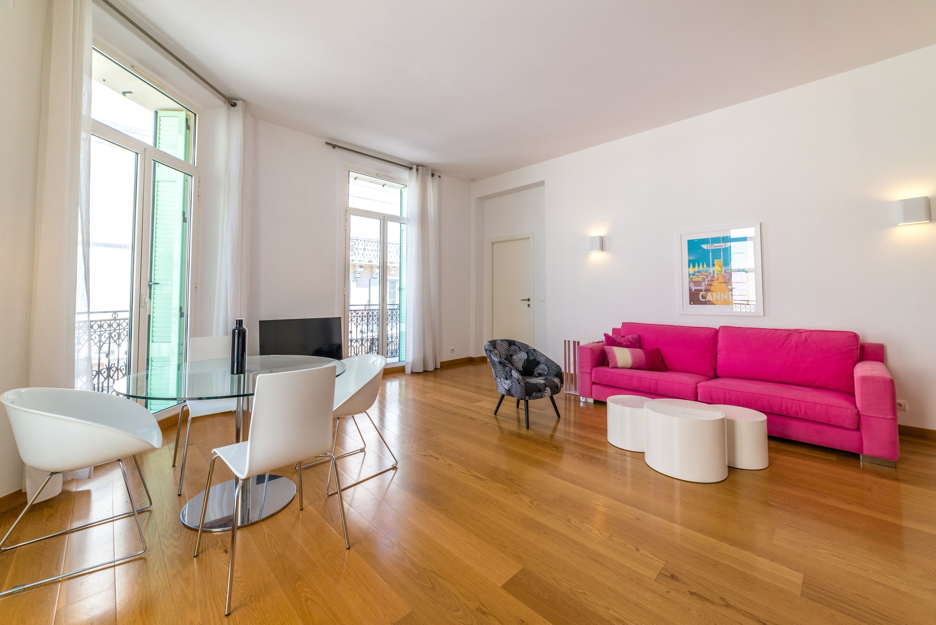 Uma sala de estar moderna com uma mesa de jantar de vidro, cadeiras brancas, uma poltrona com padrão preto e branco, um sofá rosa e um cartaz na parede.