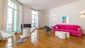 Een moderne woonkamer met een glazen eettafel, witte stoelen, een zwart-wit gestreepte fauteuil, een roze bank en een poster aan de muur.