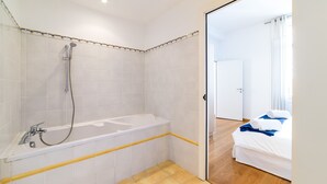 Een badkamer met bad, douche en toilet. In de aangrenzende kamer ligt een houten vloer en staat een bed met handdoeken.