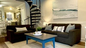 Un salon moderne avec une table basse bleue, deux canapés, un escalier en colimaçon et un grand tableau sur le thème de l’océan accroché au mur.