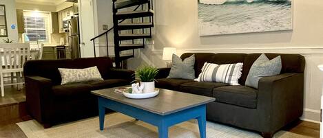 Een moderne woonkamer met een blauwe salontafel, twee banken, een spiltrap en een groot schilderij met een zeemotief aan de muur.