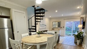 Un coin repas avec une table en bois et des chaises blanches, un escalier en colimaçon, une cuisine dotée d’un réfrigérateur en acier inoxydable et une vue sur l’extérieur, visible à travers une porte vitrée.