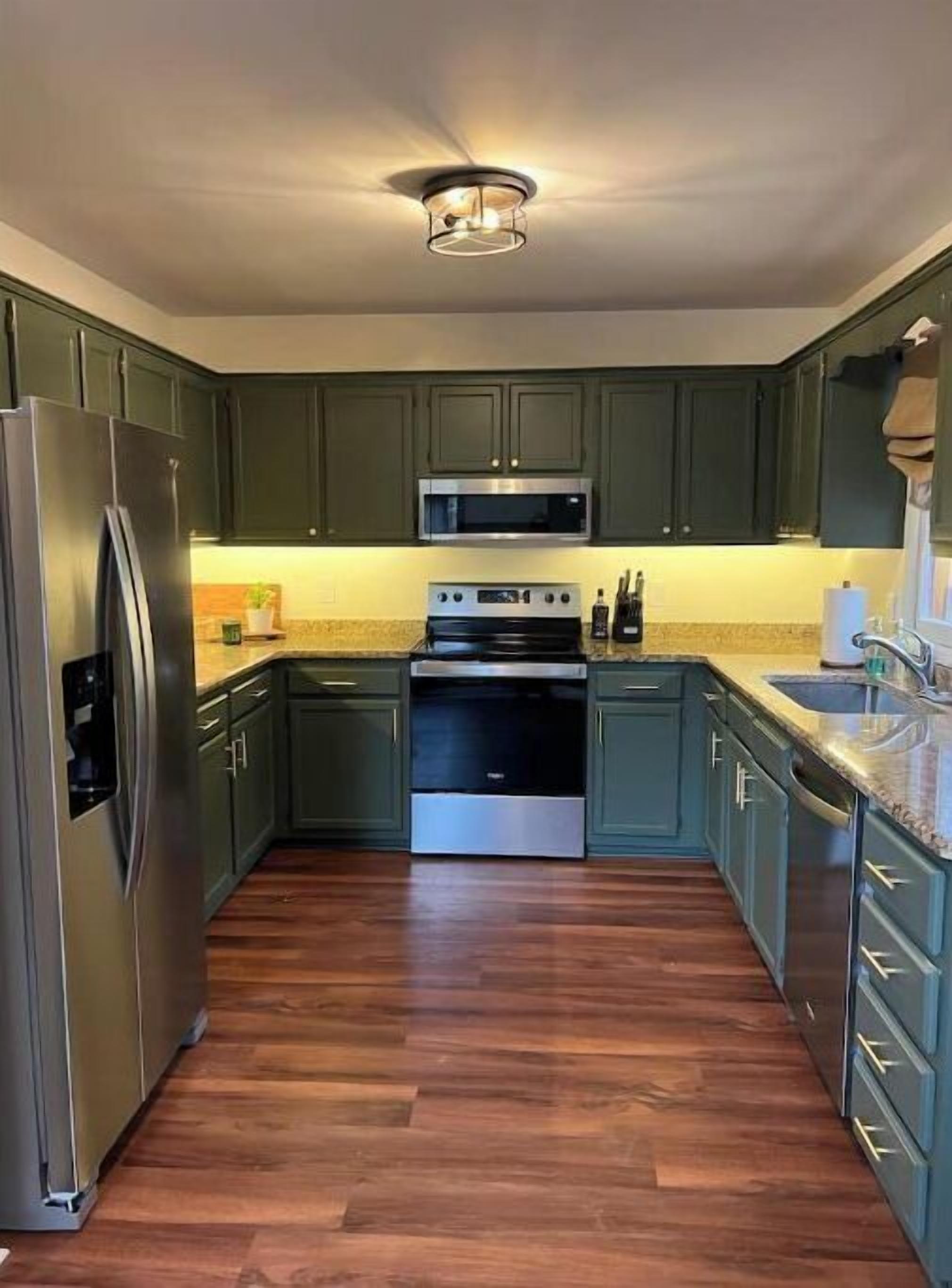 A kitchen with dark green cabinets, stainless steel appliances, and a wooden floor.