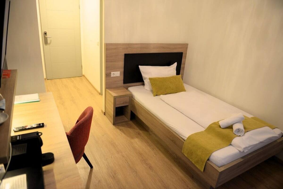 A single bed with a wooden headboard, a bedside table, a chair, and a desk with a remote control.