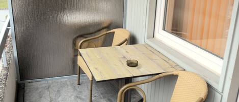 A small outdoor patio with a table and two chairs, a window with blinds, and a textured wall.