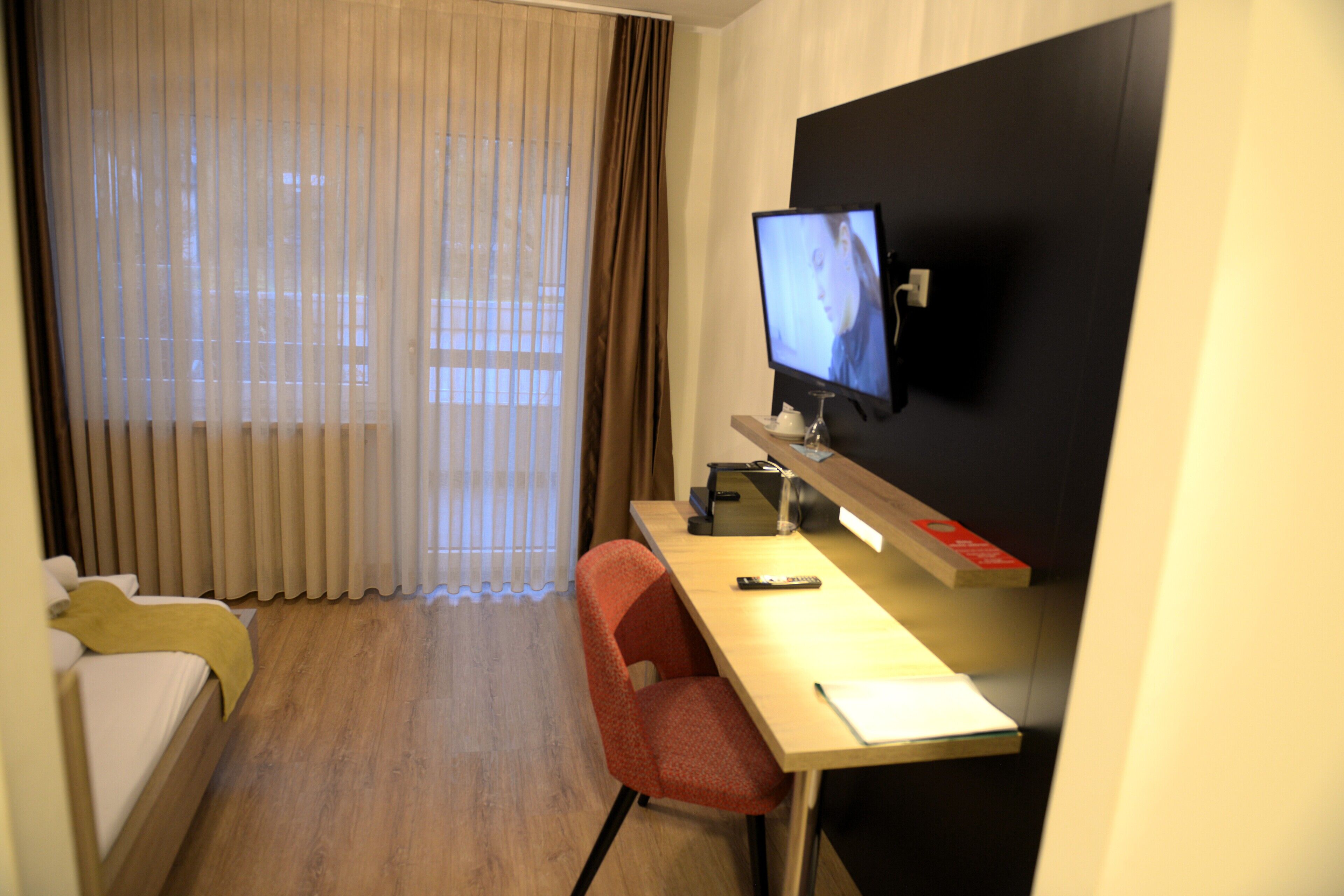 Une chambre d’hôtel avec un lit, un bureau avec un ordinateur, une télévision et une fenêtre avec des rideaux.