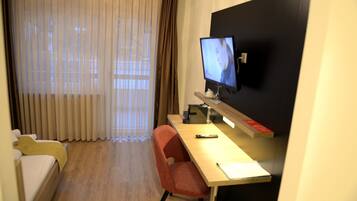 Une chambre d’hôtel avec un lit, un bureau avec un ordinateur, une télévision et une fenêtre avec des rideaux.