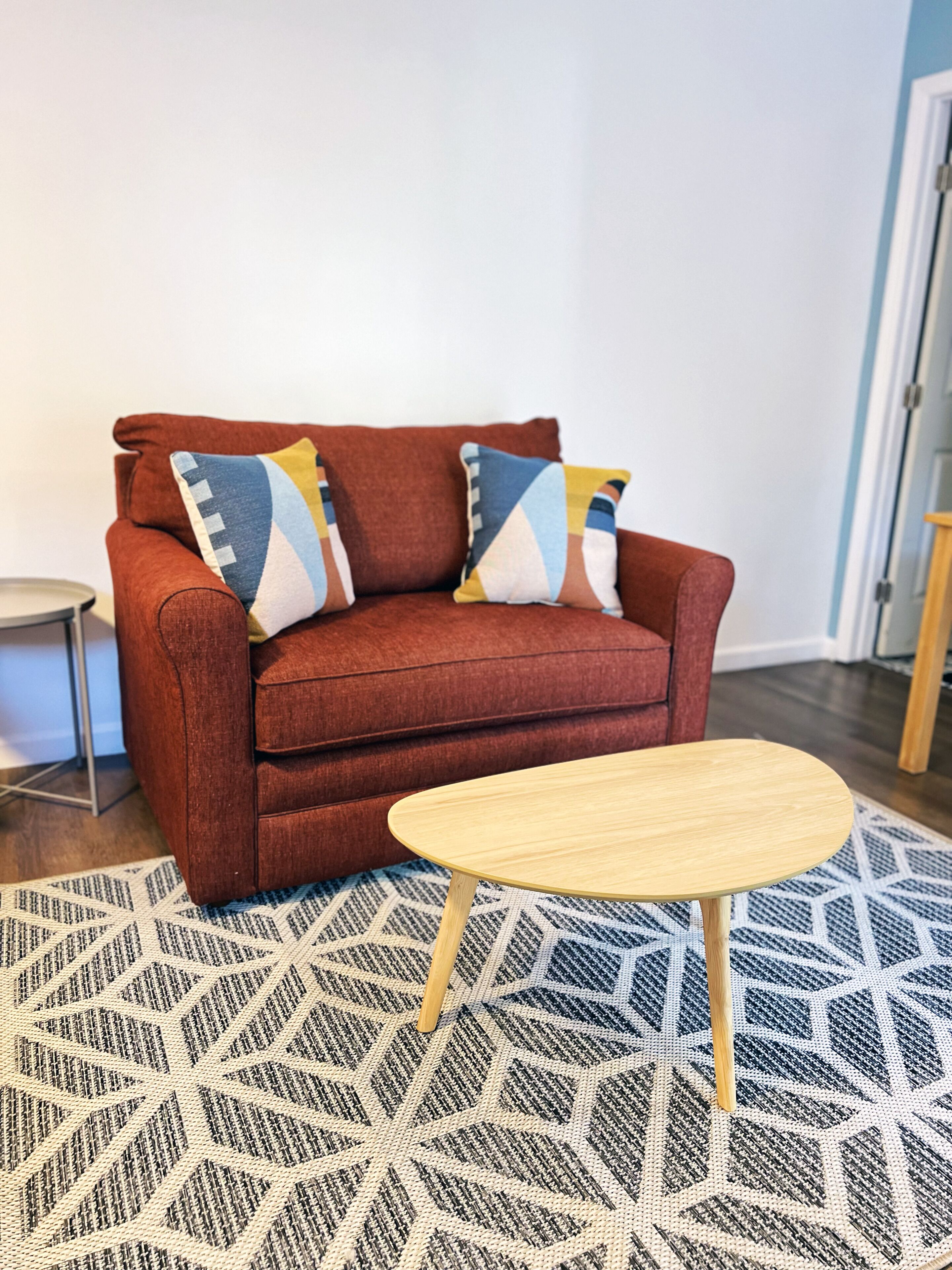A red sofa with patterned cushions, a small wooden side table, and a geometric patterned rug.