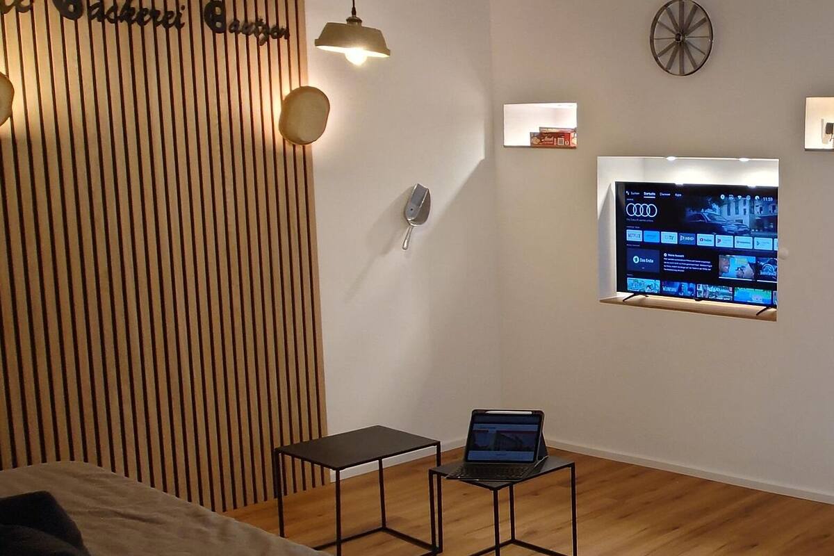 Ein modernes Zimmer mit einer Wand aus Holzlatten, einem kleinen Schreibtisch mit einem Laptop, einem an der Wand befestigten Fernseher und einer Tür mit Spiegel.