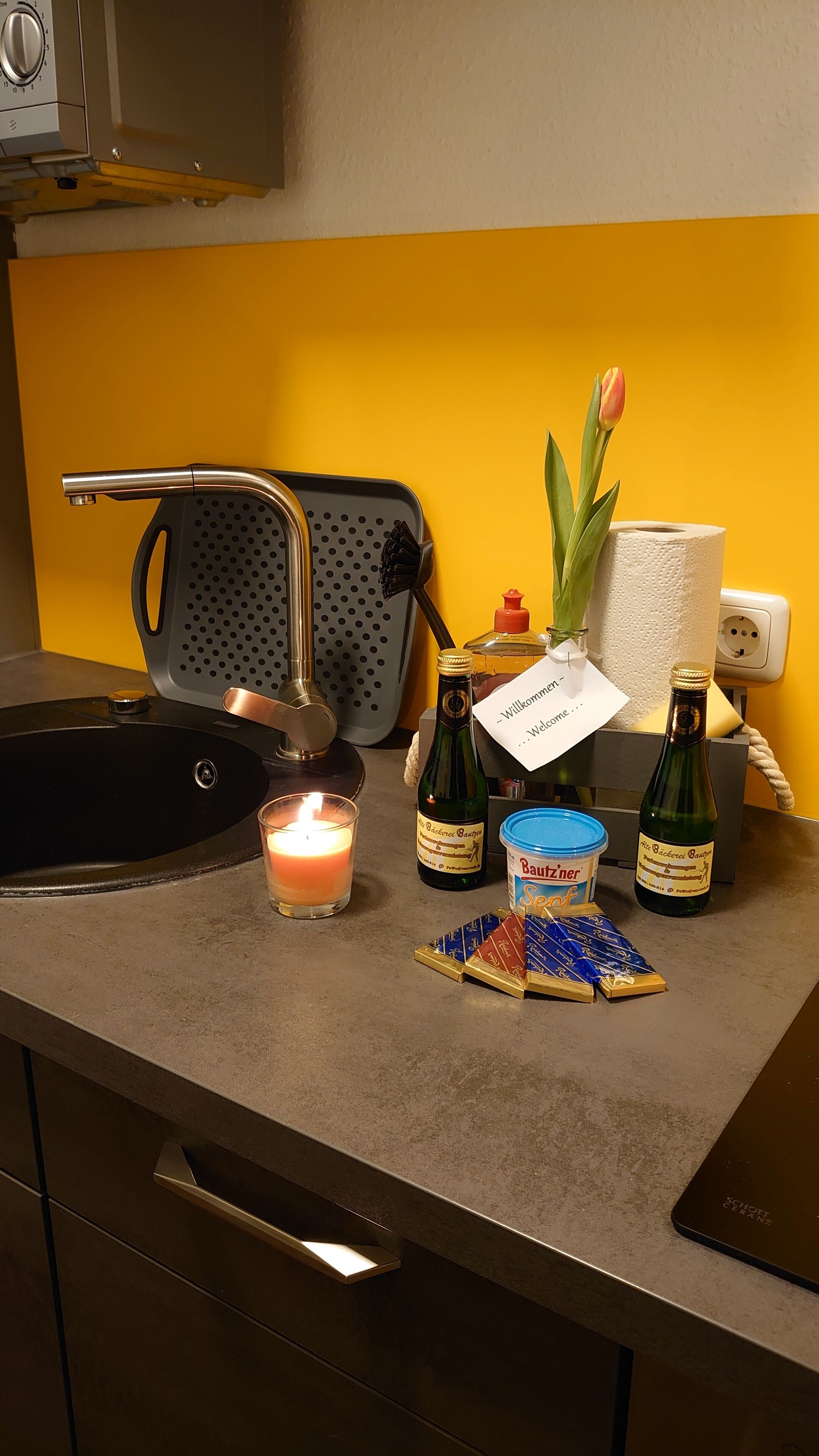 Una encimera de cocina con un lavamanos, una vela, dos botellas de cerveza, un portalapapeles y una tarjeta de bienvenida.
