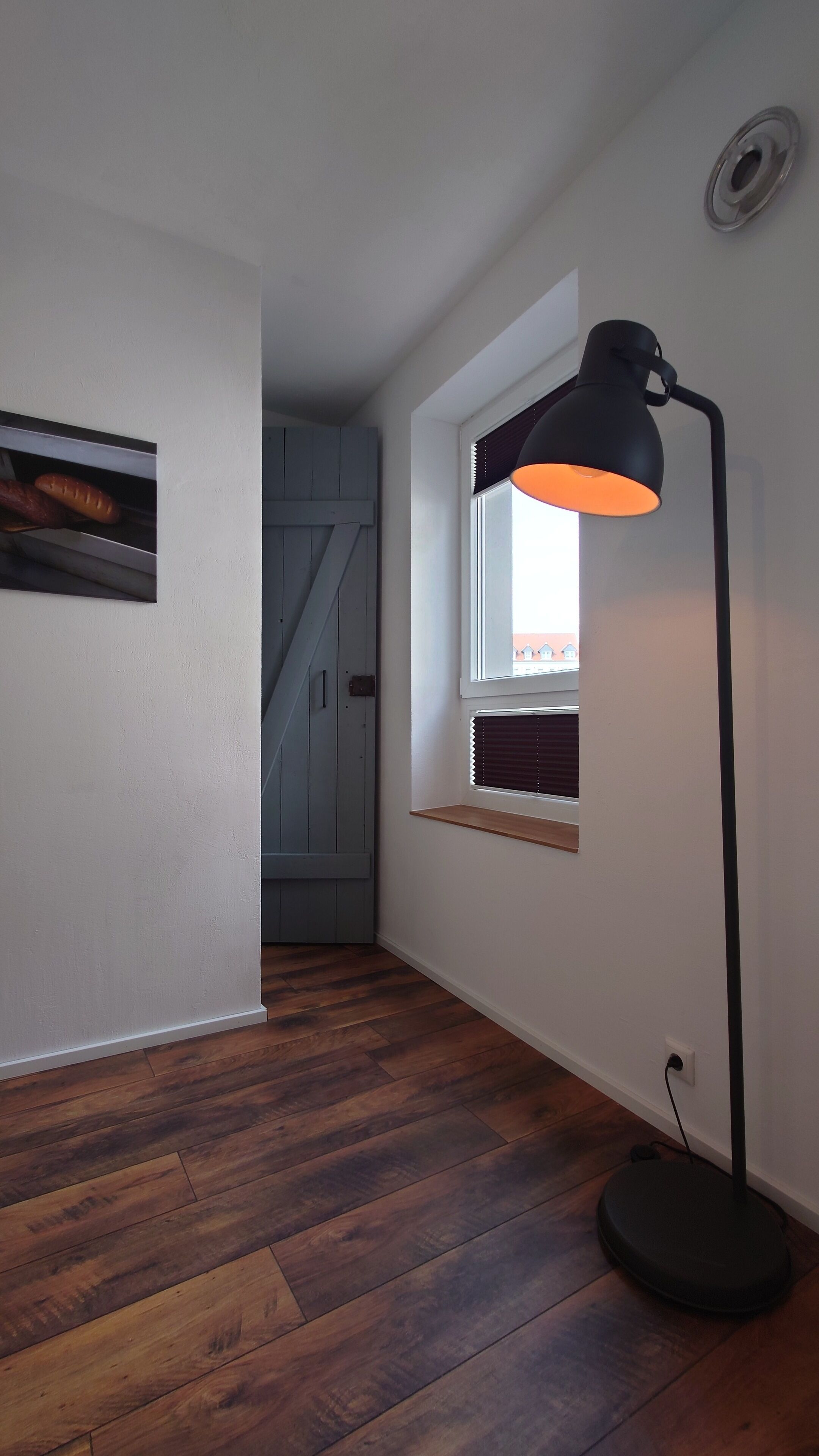 A hallway with wooden flooring, a black floor lamp, a window with blinds, and a door leading to another room.