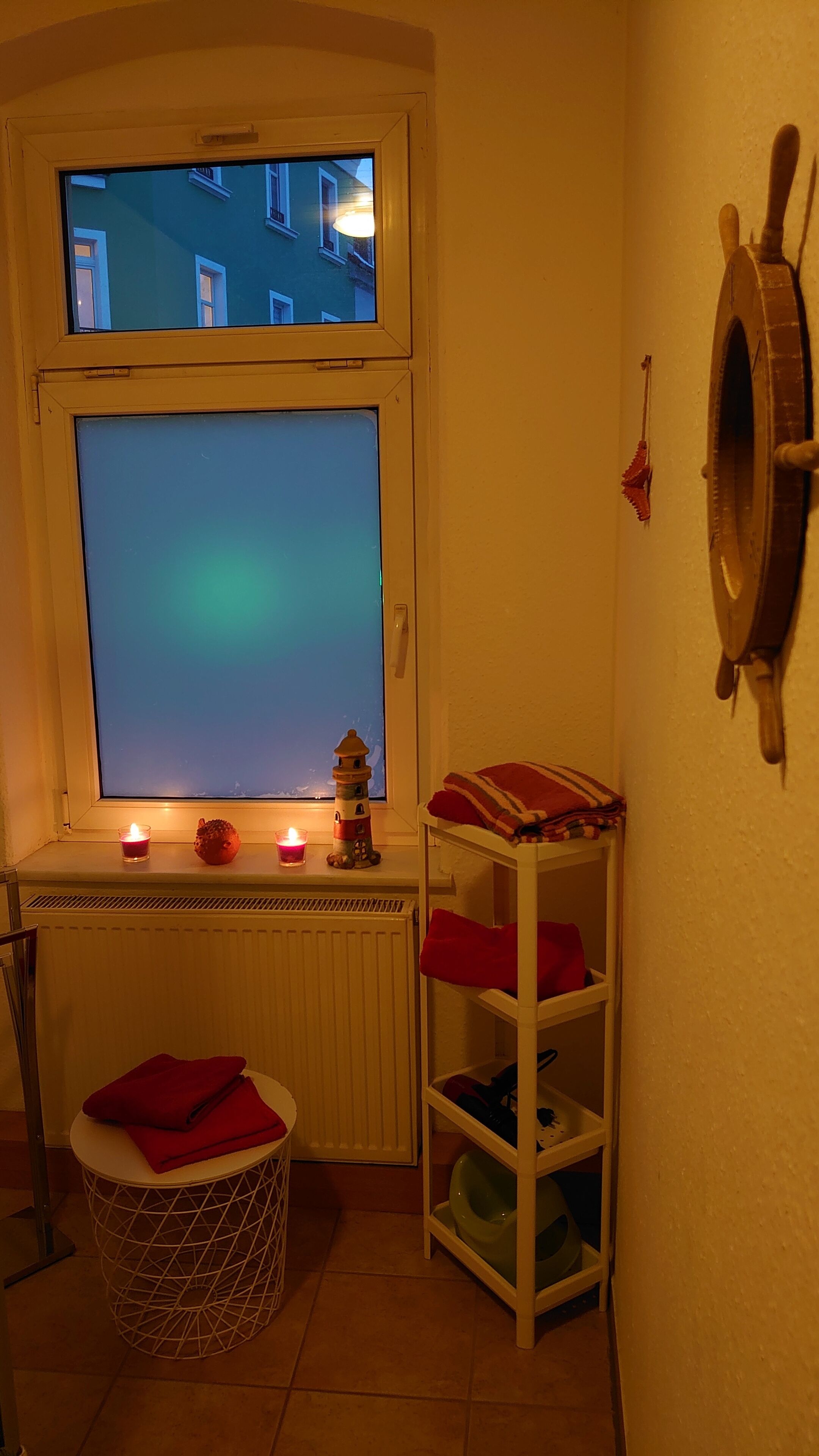 Una stanza d'angolo con un'ampia finestra, un termosifone, un tavolino con una tovaglia rossa, uno scaffale con una tovaglia rossa e un oggetto fissato al muro.