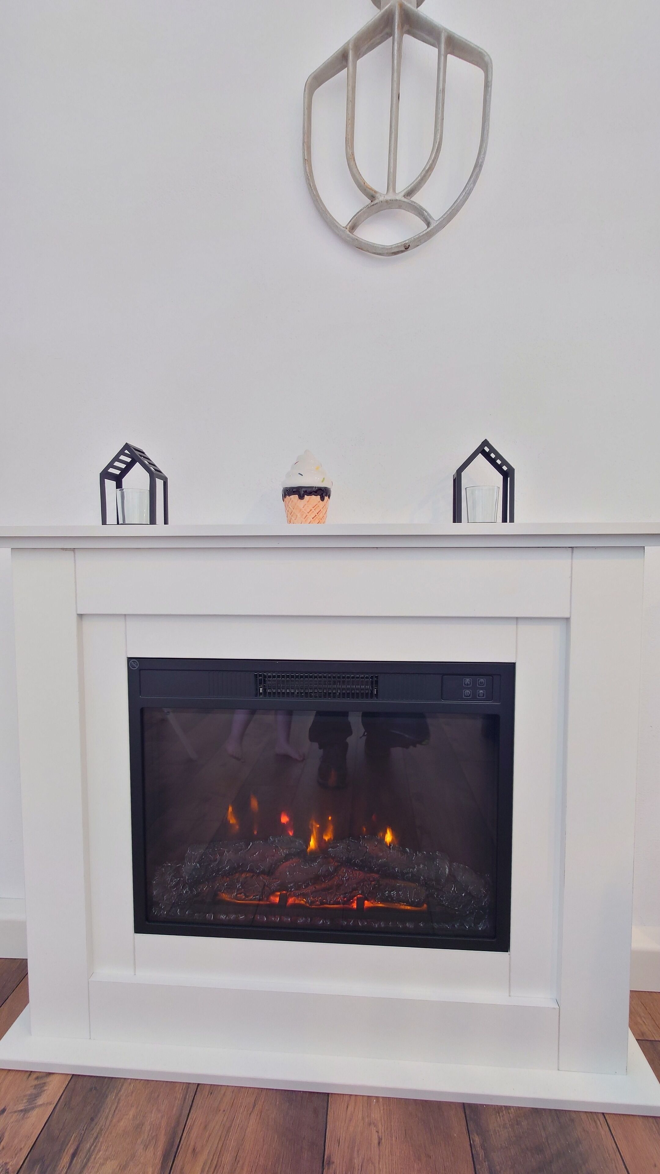 Una chimenea blanca con frente de vidrio, una pieza decorativa de metal encima y dos pequeños objetos con forma de casa en el repisa.