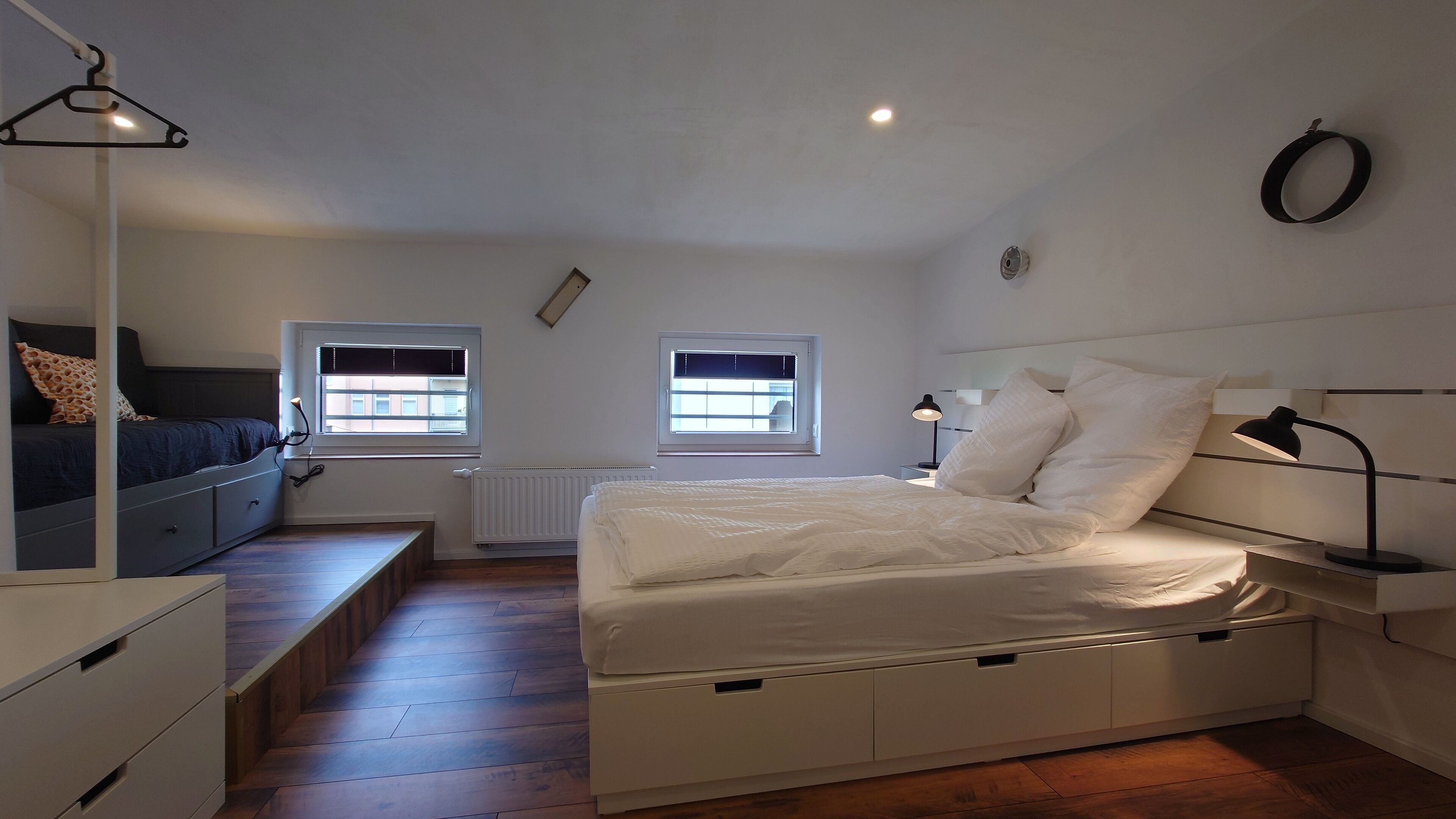 A bedroom with a bed, bedside tables, a desk, two windows, and a radiator.